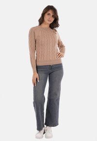 Maglione beige a trecce con maniche lunghe e scollatura rotonda, abbinato a jeans svasati a vita media e sneakers bianche. Texture liscia.