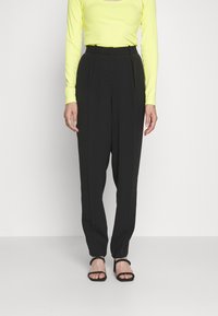 Mustat räätälöidyt housut korkealla vyötäröllä, laskostettu etuosa ja sileä pinta. Yhdistetty pitkähihainen kirkkaan keltainen paita ja mustat sandaalit.