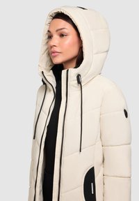 Béžová bunda s výplní, se zapínáním na zip, černými šňůrkami, oversized kapucí a boční kapsou s texturovaným akcentem.