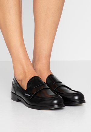 Stopy w czarnych, skórzanych loafersach Calvin Klein na niskim obcasie, na jednolitym jasnym tle.