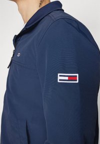 Tamnoplava jakna s ovratnikom na zip, teksturiranom tkaninom i logotipom s crvenim, bijelim i plavim prugama na lijevom rukavu.