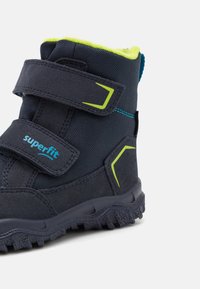 Csizma sötétkék textil felsőrésszel, zöld részletekkel, tépőzáras rögzítéssel és strapabíró fekete gumitalppal a tapadás érdekében.