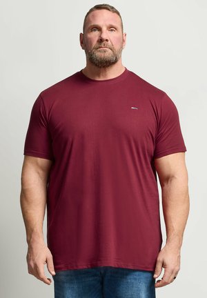 Homme musclé avec une barbe portant un t-shirt bordeaux uni à manches courtes et un jean bleu, debout sur un fond gris clair.