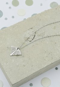 Silverhalsband med ett fläktformat hänge med linjära detaljer. Positionerat på en texturerad grå stenyta, omgiven av cirkulär dekoration.