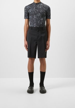 Černé plisované šortky spojené s krátkým rukávem a tmavým vzorovaným topem. Černé ponožky a nazouvací boty doplňují outfit, zdůrazňující moderní vzhled.