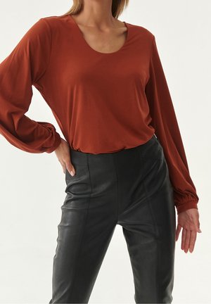 Femme portant un chemisier à manches longues couleur rouille rentré dans un pantalon en cuir noir taille haute, posant avec une main sur la hanche devant un fond blanc.