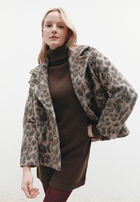 Leopardenmuster-Wollmischungsmantel mit Reverskragen, zwei Knöpfen und einem strukturierten braunen Rollkragenpullover darunter. Dunkel vertikal gerippte Strumpfhosen.