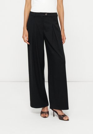 Femme portant un pantalon noir à jambes larges avec des plis sur le devant, associé à des sandales à talons ouvertes foncées et un haut blanc.