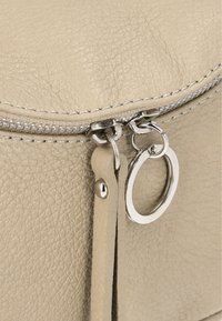 Beige lædertaske med en tekstureret overflade, prydet med en sølvfarvet lynlås og metalringe, detaljeret med kontrasterende syninger.
