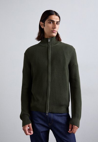 Joven con cabello largo y oscuro, vestido con un suéter de cremallera de canalé en color verde oliva y jeans oscuros, de pie frente a un fondo liso.