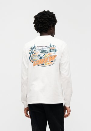 Bela majica z dolgimi rokavi z barvno grafiko na hrbtni strani, ki prikazuje karirane zastave, besedilo in plamene. Izdelana iz bombažnega blaga.