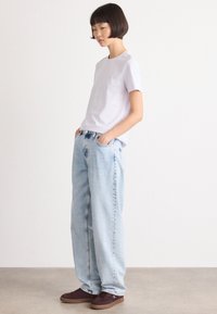 Lys lilla t-skjorte, kort erm, kombinert med løstsittende, falmede blå jeans. Modellen har brune joggesko, hendene i lommene, står på siden.