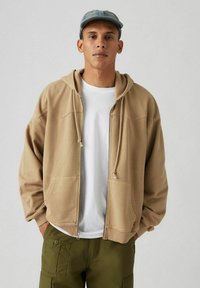 Beige zip-up hoodie met een trekkoordkap, kangoeroe pockets en stiksel details, gecombineerd met een wit T-shirt en olijfgroene broeken.