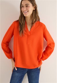 Femme souriante, les mains sur les hanches, portant un chemisier à manches longues orange vif et un jean bleu sur un fond neutre.
