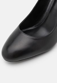 Chaussure à talon haut en cuir noir avec un bout arrondi et une texture lisse. Le design présente une tige basse et un minimum de coutures apparentes.