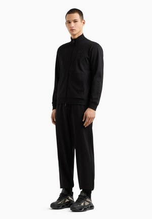 Giacca nera con zip e colletto alto, abbinata a pantaloni neri larghi. Le sneakers presentano dettagli metallici. Tessuto morbido e liscio.