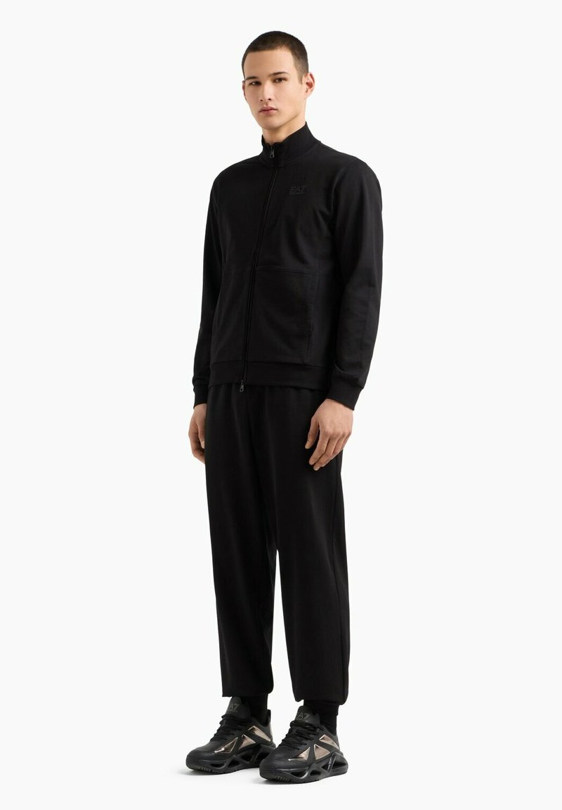 Giacca nera con zip e colletto alto, abbinata a pantaloni neri larghi. Le sneakers presentano dettagli metallici. Tessuto morbido e liscio.