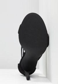 Sandale noire à talon haut positionnée semelle vers le haut contre un coin blanc, montrant un talon étroit et une surface de semelle texturée.