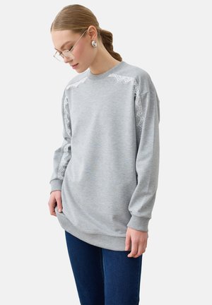 Kvinde med briller iført en lysegrå sweatshirt med lange ærmer og blonde detaljer på skuldre og ærmer, kombineret med mørkeblå jeans.