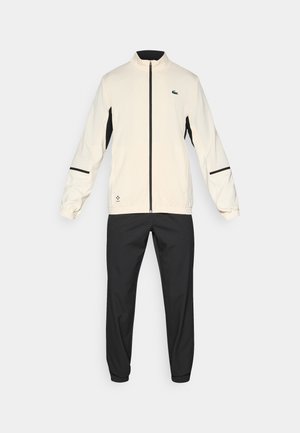 Track suit featuring a cream jacket with black accents and zip pockets, paired with black pants with elastic cuffs. Smooth fabric texture.