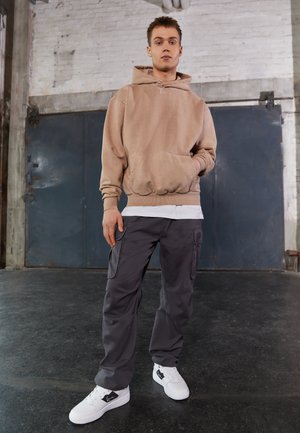 Beige hettegenser med lommer foran, sammen med mørkegrå cargo-bukser med sidelommer, og hvite sneakers med svarte detaljer.