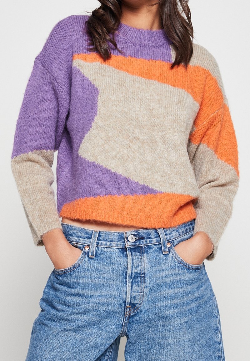 Maglione a maglia in viola, arancione e beige con motivi geometrici. Presenta un collo rotondo e una vestibilità ampia. Abbinato a jeans a vita alta.