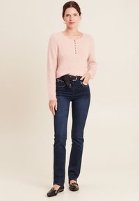 Weicher, rosa flauschiger Pullover mit langen Ärmeln und Rundhalsausschnitt, ausgestattet mit einem Knopfdetail. Kombiniert mit dunkelblauen Slim-Jeans und schwarzen Ballerinas.
