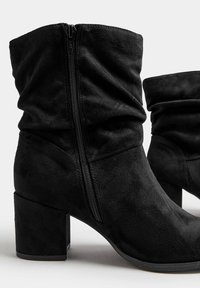 Botines de ante negro con un diseño acampanado, que cuentan con una cremallera lateral y un tacón grueso. Textura suave y puntera redondeada.
