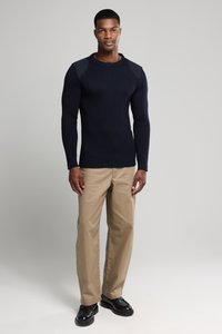 Granatowy sweter z prążkowanej tkaniny z akcentami na ramionach, zestawiony z beżowymi spodniami i czarnymi butami. Tkanina wydaje się miękka, a krój jest dopasowany.