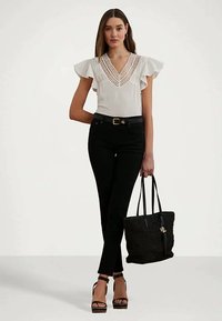 Frau, die eine weiße Bluse mit Spitze und schwarze Hosen trägt, eine schwarze geflochtene Tasche hält und vor einem weißen Hintergrund steht.