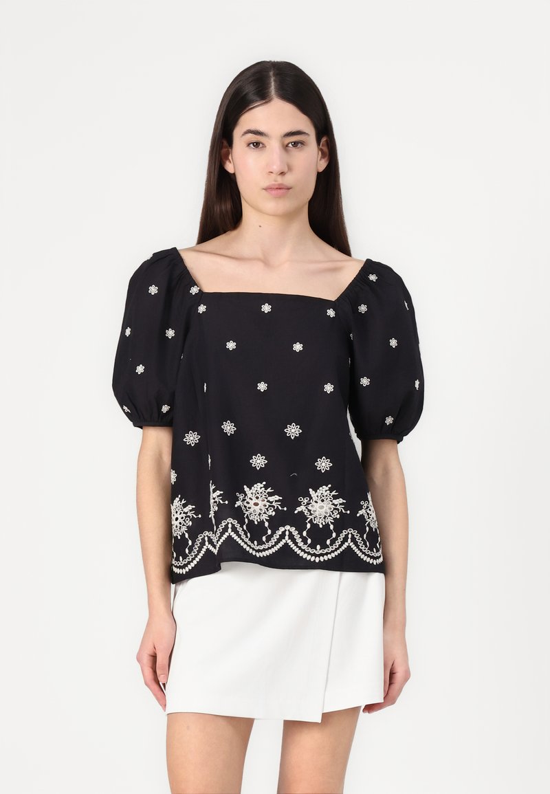 Top nero a maniche puff con scollatura quadrata, adornato con ricami floreali bianchi e rifiniture decorative sull'orlo. Indossato con una gonna bianca.
