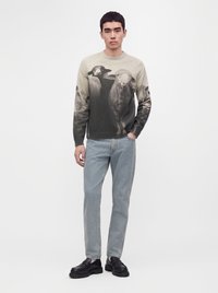 Jeune homme portant un pull avec un motif de moutons noir et blanc, un jean bleu clair et des chaussures noires, debout devant un fond uni.