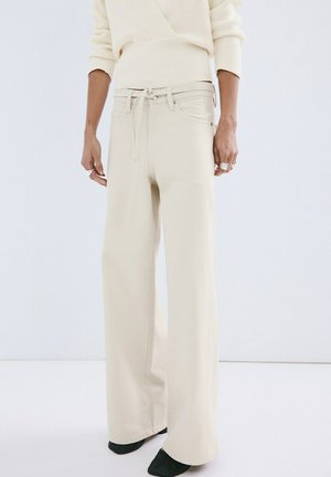 Persona con pantalones anchos color crema con cintura ajustable y un suéter de punto color crema, de pie frente a un fondo liso.