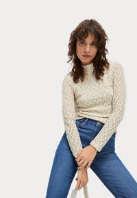 Top lungo in pizzo crema con maniche a sbuffo e collo alto, caratterizzato da motivi floreali. Abbinato a jeans in denim blu. Modella che tiene una borsa chiara.