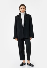 Zwarte oversized blazer met notch-kleppen, enkele knoopsluiting, gecombineerd met zwarte op maat gemaakte broek en glanzende zwarte instappers. Wit coltrui eronder.