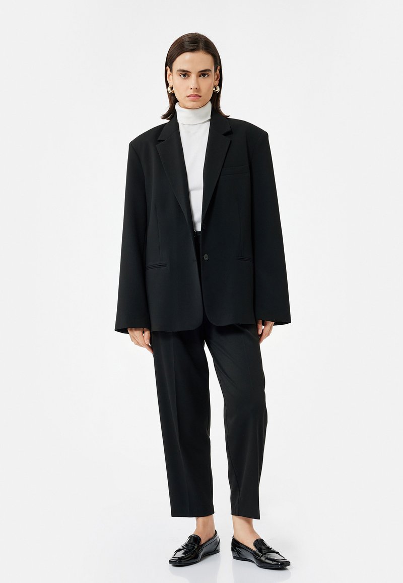 Zwarte oversized blazer met notch-kleppen, enkele knoopsluiting, gecombineerd met zwarte op maat gemaakte broek en glanzende zwarte instappers. Wit coltrui eronder.