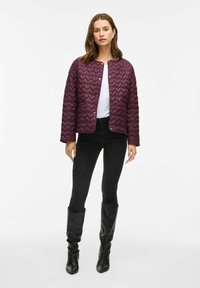 Bordeaux quilts jasje met een ronde halslijn, drukknoopjes en een gestructureerd patroon, gecombineerd met zwarte slim-fit jeans en zwarte laarzen tot de knie.