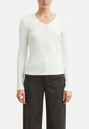 Frau trägt ein weißes, geripptes Langarmshirt mit V-Ausschnitt und locker sitzende dunkle Hose und steht vor einem schlichten Hintergrund.