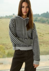 Szara i czarna bluza z kapturem w paski z przednią kieszenią i troczkami, połączona z ciemnobrązowymi spodniami; luźny krój, miękka faktura.