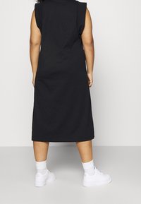 Czarna bezrękawnikowa sukienka midi z opadającymi ramionami, wykonana z bawełny. Połączona z białymi sneakerami i skarpetkami sięgającymi kostek, na jasnym tle.