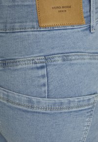 Os jeans de denim azul claro apresentam um remendo de couro castanho-claro na parte de trás, com as inscrições "VERO MODA" e "DENIM" em relevo, além de costuras visíveis e um design de bolso.
