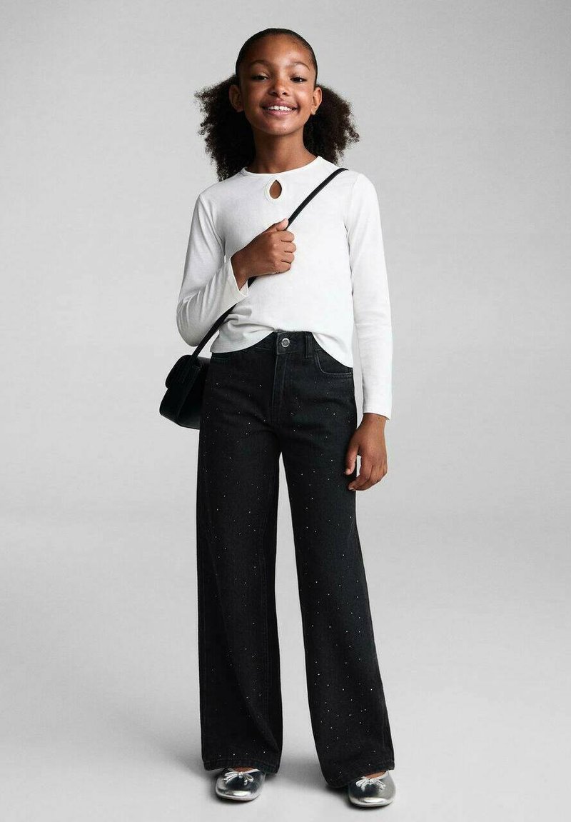 Černé široké džíny se stříbrnými ozdobami, spárované s bílým topem s dlouhým rukávem a výřezem ve tvaru klíčové dírky, a černou crossbody taškou.