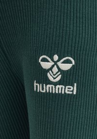 Grön ribbad tyg med en broderad vit logotyp som har en stiliserad bi och texten "hummel" nedanför designen.
