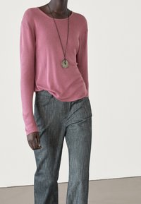 Roze gebreide long-sleeve top gecombineerd met grijze, gestructureerde broeken. Het model draagt een eenvoudige ronde hangerketting en toont een ontspannen pasvorm.