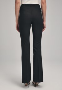 Zwarte flared broek van een gladde stof, met een op maat gemaakte tailleband en minimale steken. Draagt met zwarte hoge hakken.