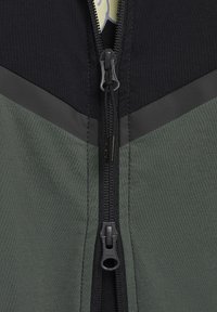 Chaqueta con cremallera que presenta una sección superior negra, una sección inferior verde texturizada y un diseño en forma de chevrón gris. Incluye una cremallera resistente.
