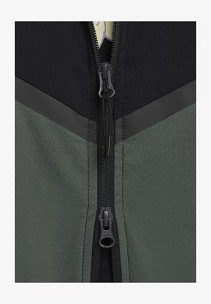 Chaqueta con cremallera que presenta una sección superior negra, una sección inferior verde texturizada y un diseño en forma de chevrón gris. Incluye una cremallera resistente.