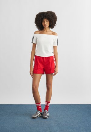 Naine seisab sinisel vaibal, kandes valget õlgadeta pluusi, punaseid spordipükse, punaste triipudega sokke ja hõbedasi tossusid valgel taustal.