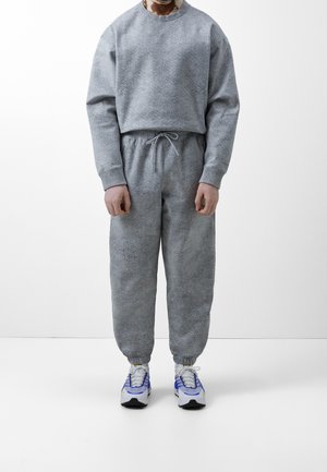 Personne portant un sweat-shirt gris clair texturé et un pantalon de jogging assorti avec des baskets blanches et bleues, debout devant un fond blanc uni.