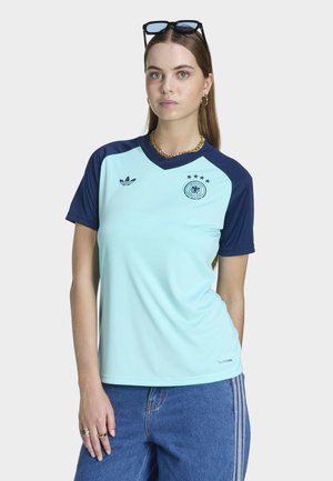 Donna che indossa una maglia da calcio celeste e blu navy con l'emblema della Germania, jeans blu, collana d'oro, orecchini a cerchio e occhiali da sole in testa.
